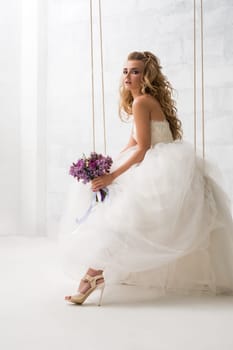 Young beautiful bride with gorgeous hair style posing on a swing holding nice bouquet studio shot