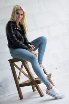 Sexy blonde in black leather jaket and blue jeans sitting on the wooden chair studio portrait