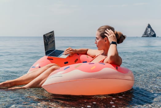 A woman in a bikini sits on a pink floatie in the ocean, working on a laptop.