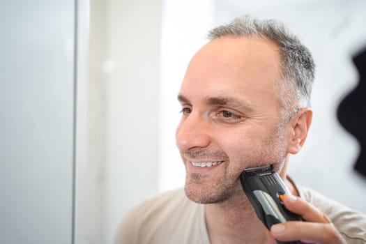 Man shaves his beard with a trimmer in the bathroom.