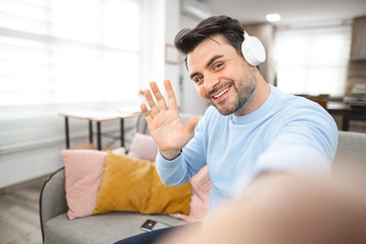 Brown-haired man with white headphones on his head sits on the couch in the living room and makes a selfie on a mobile phone.