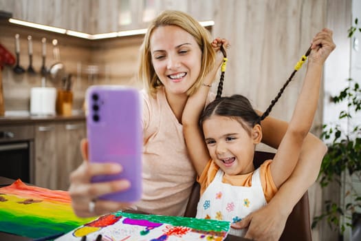 A mother and daughter enjoy a playful selfie moment while surrounded by colorful drawings and crafts in their cozy kitchen.