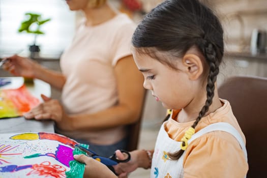 A child focused on cutting her colorful drawing with scissors, while an adult works on a similar project in the background.