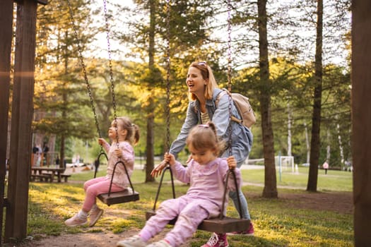 Mother is rocking her twin daughters on a swing in the park.