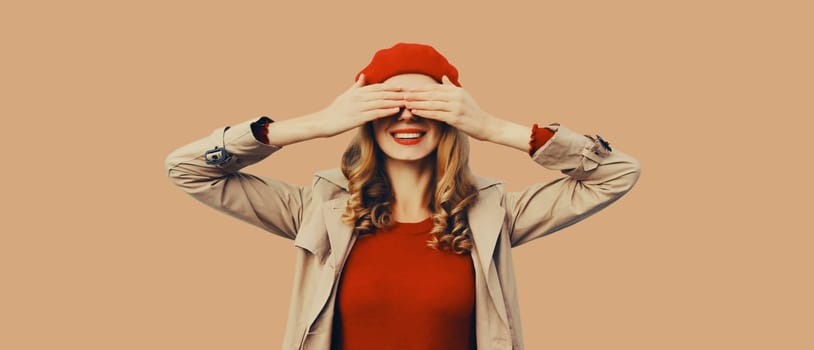 Stylish surprised young woman covering her eyes with hands, fashionable girl in red french beret hat, jacket posing on brown background