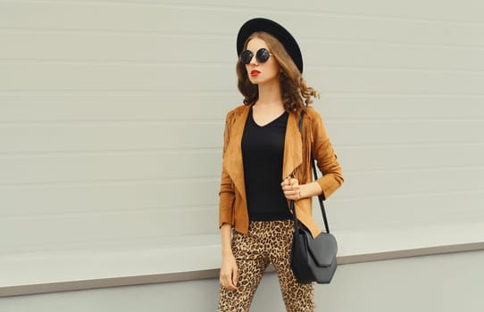Beautiful stylish young woman posing with handbag looking away in black hat, jacket, glasses on city street on gray background