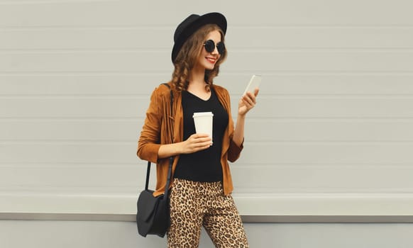 Stylish happy smiling young woman with phone, beautiful girl holding smartphone, cup of coffee wearing hat standing on city street on gray background