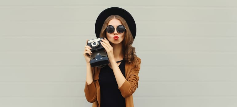 Portrait of beautiful happy young woman photographer taking picture with camera, stylish creative girl in black hat on gray background