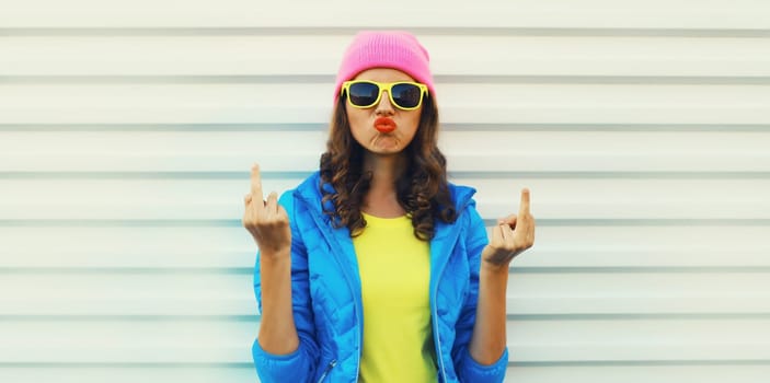 Portrait of bad angry young woman expression showing hand with middle finger sign, stylish girl wearing vivid colorful clothing, pink hat, glasses on white background