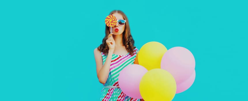 Beautiful young woman with bunch of balloons blowing kiss in colorful dress on blue background