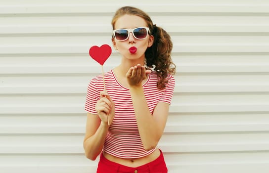 Beautiful young woman with red heart shaped sunglasses blowing kiss on white wall background