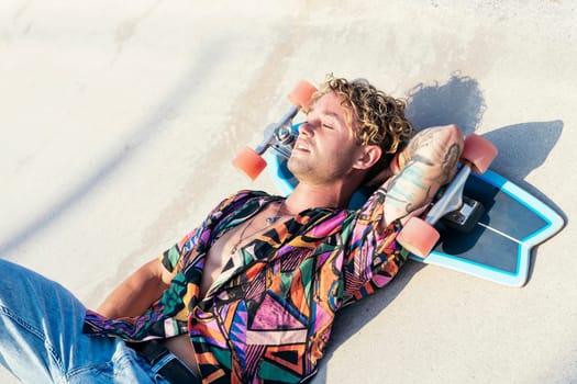 high angle of young hipster tattooed male in stylish colorful shirt lying on concrete ramp with skateboard under head and enjoying summertime in skatepark