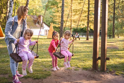 Mother is rocking her twin daughters on a swing in the park.