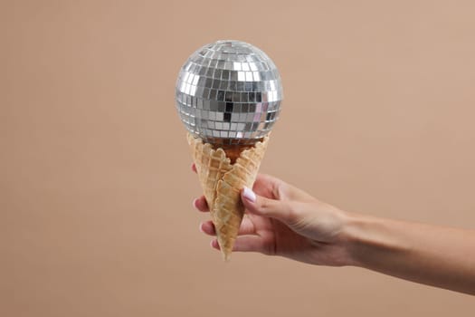 Hand holding waffle cone with disco ball on top, on neutral beige background. Disco party concept.
