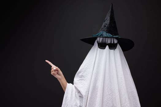 Person in costume of ghost pointing at something on dark background with space for text. Halloween celebration