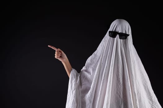 Person in costume of ghost pointing at something on dark background with space for text. Halloween celebration