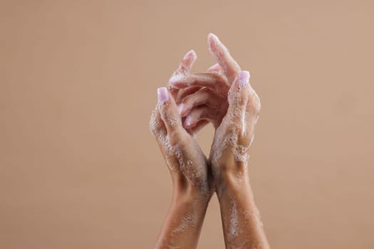 A close-up of two hands lathered with soap, demonstrating hand washing. The hands are positioned in a way that emphasizes the soap bubbles against a neutral brown background.