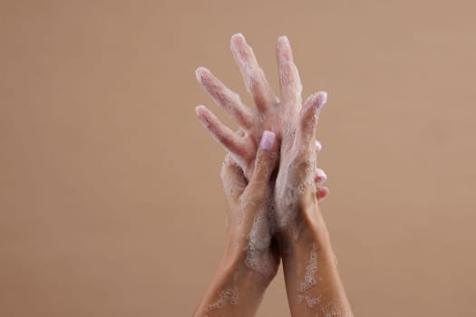 A close-up of two hands lathered with soap, demonstrating hand washing. The hands are positioned in a way that emphasizes the soap bubbles against a neutral brown background.