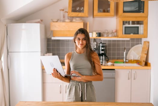 A young woman in a modern kitchen confidently holds a sleek laptop, embodying remote work and productivity