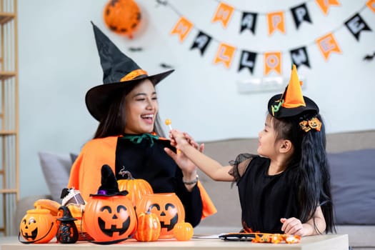 Playful young mother sitting on comfy couch in living room with cute little daughter tells spooky Halloween stories. Funny mom and girl kid have fun at home. Celebration and Autumn holiday concept.