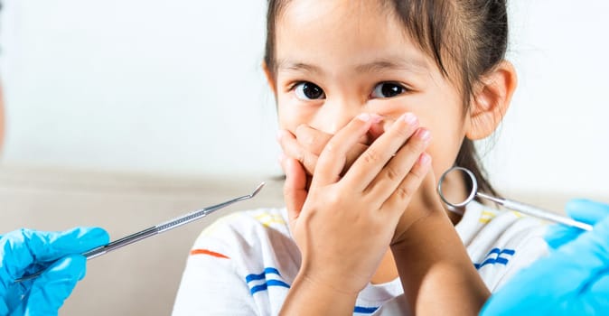 Dental kid examination. Doctor examines oral cavity of child uses mouth mirror to check teeth cavity but little girl cover mouth no need to checking she afraid, scared and closes his mouth with hands