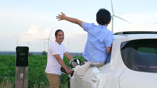 Concept of progressive happy family enjoying their time at wind farm with electric vehicle. Electric vehicle driven by clean renewable energy from wind turbine generator for charging station.