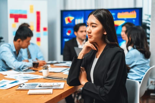 Portrait of happy young asian businesswoman or analyst looking at camera with her colleague analyzing data analysis in dynamic business strategy investment planning meeting. Habiliment