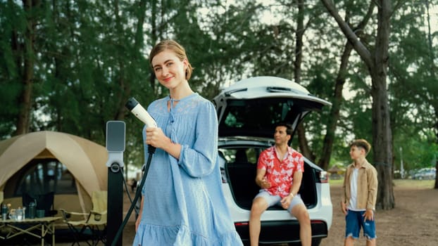 Outdoor adventure and family vacation camping at sea travel by eco friendly car. Cheerful woman or mother holding, pointing EV charger point with playful and happiness posture in campsite. Perpetual