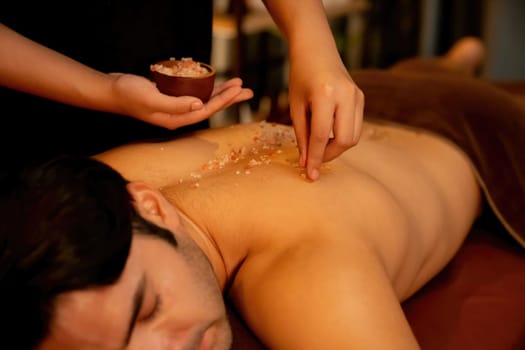 Man customer having exfoliation treatment in luxury spa salon with warmth candle light ambient. Salt scrub beauty treatment in Health spa body scrub. Quiescent