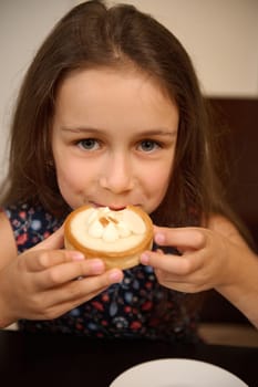Close-up portrait of a Caucasian cute little child girl tasting a sweet French baked dessert - a lemon tartlet, smiles looking at camera