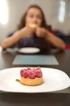 Close-up of a tasty sweet French dessert - a tartlet with raspberries on white plate, over blurred background of a little child taking her breakfast or snacking. Food still life. Culinary. Patisserie