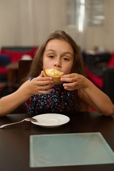 Authentic portrait of a cute little child girl sitting at table and enjoying eating fresh sweet sugary dessert - a tartlet with lemon according to traditional French recipe. Food and bakery concept