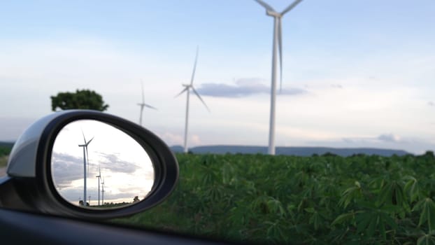 Progressive future energy infrastructure concept of wind turbine reflected in side mirror of electric vehicle being charged at charging station powered by green and renewable energy from wind turbine.