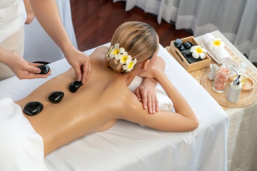 Hot stone massage at spa salon in luxury resort with day light serenity ambient, blissful woman customer enjoying spa basalt stone massage glide over body with soothing warmth. Quiescent
