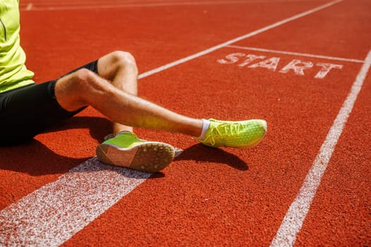 A close-up of a runner's leg and foot on a red athletic track, positioned near the starting line marked with the word START. The runner is wearing bright yellow shoes and black shorts