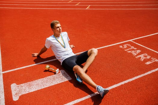 A young male athlete sitting on a running track, smiling and relaxed. He is wearing a white t-shirt and black shorts, with a medal around his neck and a trophy beside him