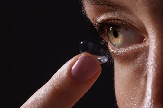 A close-up of a woman applying a contact lens to her eye. The focus is on her eye and the lens, showcasing the delicate process of lens application. The background is dark, emphasizing the subject.