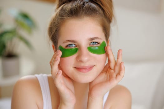 Young woman with green under-eye patches, smiling and touching her face.