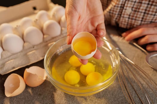 A person cracking an egg into a glass bowl, with several eggs in an egg carton in the background. The scene is set in a kitchen, showcasing the process of preparing food.