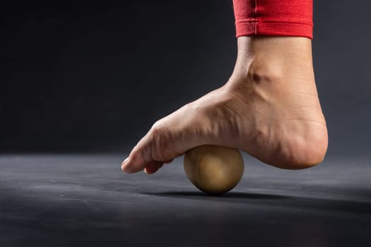 Myofascial release of foot muscles with a massage ball on a dark background, close-up. Concept on the topic of preventing foot fatigue after wearing high-heeled shoes.
