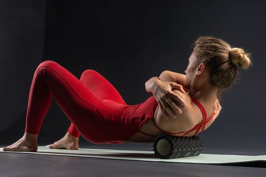 Woman performing myofascial release of hyperelastic back muscles with a massage roller on a dark background. Concept of back fatigue prevention and self-massage.