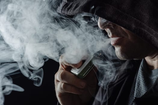 Man with vaping mod exhaling steam at black studio background. Bearded guy smoking e-cigarette to quit tobacco. Vapor and alternative nicotine free smoking concept, closeup