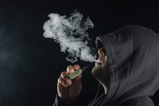 Man with vaping mod exhaling steam at black studio background. Bearded guy smoking e-cigarette to quit tobacco. Vapor and alternative nicotine free smoking concept, closeup