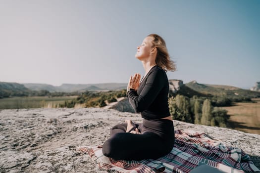A woman sits cross-legged on a mountaintop, eyes closed in a state of meditation, enjoying the natural beauty around her.