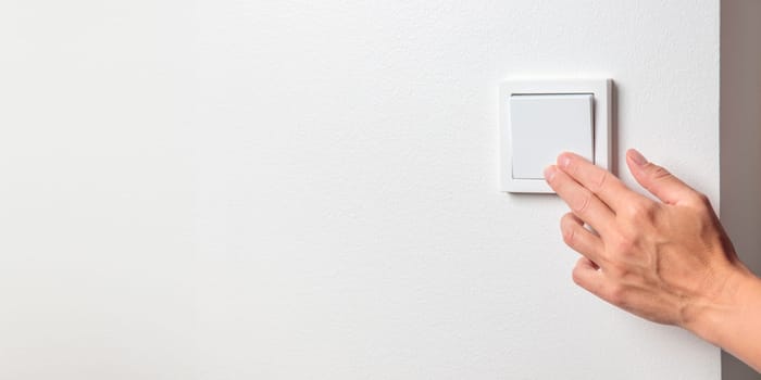 A woman's hand turning off a light switch on a white wall. The electricity conservation and mindful resource use, the impact of rising electricity costs and the need for careful energy management