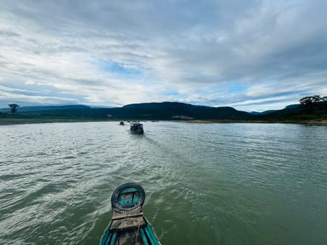Several boats navigate a calm river surrounded by lush greenery and mountains, with a cloudy sky overhead, creating a peaceful atmosphere in nature.