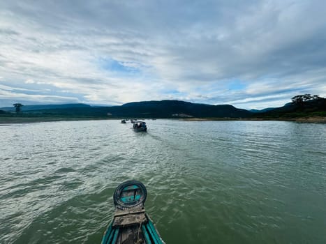 Several boats navigate a calm river surrounded by lush greenery and mountains, with a cloudy sky overhead, creating a peaceful atmosphere in nature.