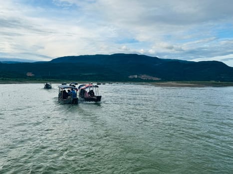 A group of boats glides smoothly across calm river waters, surrounded by towering mountains under a dramatic sky. This peaceful outing highlights nature's beauty.