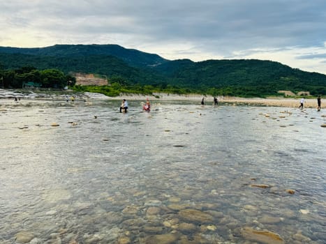 A group of individuals wades through shallow waters and rocks along the coastline, enjoying a tranquil afternoon in a picturesque natural setting with mountains in the background.