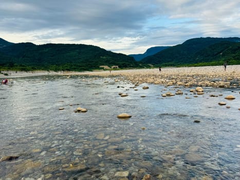 Visitors enjoy leisure time at a rocky riverbank, carefully navigating the stones while surrounded by lush mountains under a moody sky.